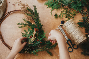 Hands holding fir branches and pine cones, berries, thread, scissors on wooden table. Making rustic...