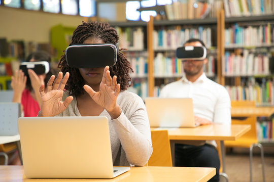 Mix raced group of students using VR experience for studying. Man and women in virtual reality headsets sitting at desks with laptops and touching air. Augmented reality concept