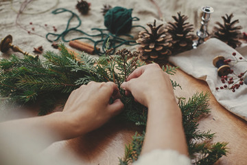 Hands holding herbs and  fir branches, pine cones, thread, berries, cinnamon on wooden table. Christmas wreath workshop. Authentic stylish still life. Making rustic Christmas wreath.