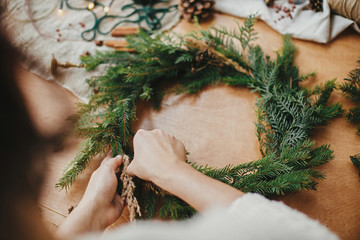 Making rustic Christmas wreath. Hands holding herbs and fir branches, pine cones, thread, berries,...
