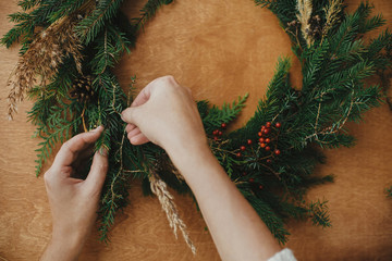 Making rustic Christmas wreath flat lay. Hands holding fir branches, and pine cones, thread, berries  on wooden table. Christmas wreath workshop. Authentic stylish still life