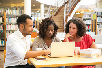 Front view of teachers working at library. Focused colleagues sitting at table with laptop. Education concept