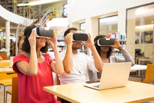 Mix raced team of adult students wearing VR goggles working on project together. Man and women in virtual reality glasses, sitting at desk with laptop. VR environment concept