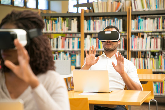 Multiethnic group of students using VR gadgets for studying. Man and woman in virtual reality headsets sitting at desks with laptops and touching air. VR simulators concept
