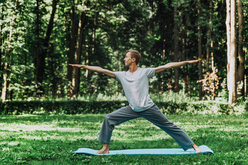 Young attractive man in a gray T-shirt and sweatpants doing yoga warrior pose in the park. He spread his legs wide, arms outstretched to the sides, looking to the side.