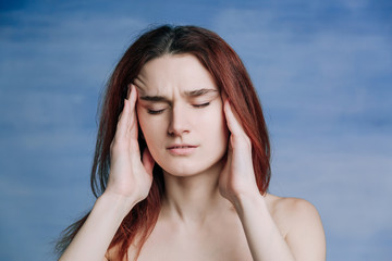 A girl with red hair twisted her face in pain and raises her hands to her face on blue background.