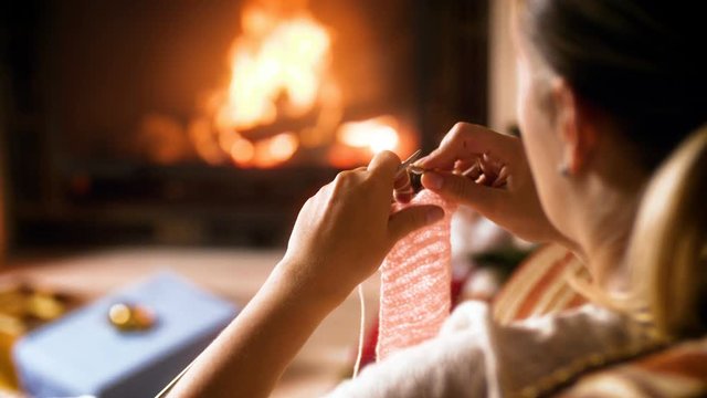 Closeup 4k footage of woman knitted woolen scarf for her children while sitting next to burning fireplace at house. People relaxing on winter holidays and celebrations at home
