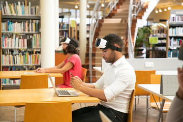 Adult students using VR technology for work on project in library. Man and woman wearing virtual reality glasses, sitting at desks, using laptop and smartphone. Innovation concept