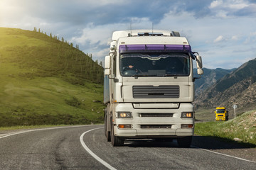 A largewhite and yellow truck rides on a highway in the mountains while transporting goods over long distances. Fast delivery by ground transportation.