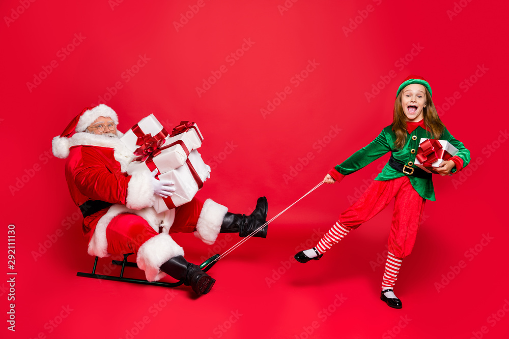 Wall mural Full size photo of funny santa claus with eyeglasses eyewear spectacles and cute elf in green hat headwear holding sledges carry sack bag with gifts isolated over red background