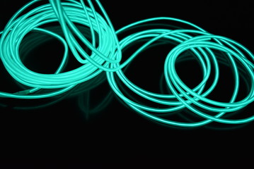 Unusual bright wires made of turquoise-colored material glowing at night. Chaotic sky blue wires, light guide electroluminescent wires, electroluminescence are located on a glossy black surface.