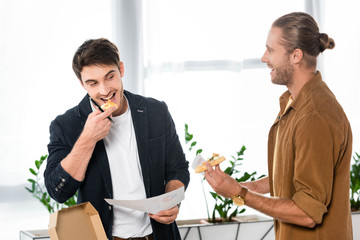 two smiling friends eating pizza and holding paper in office