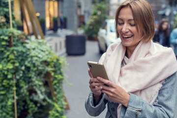 Beautiful smiling woman in a coat and a light pink scarf with a mobile phone while traveling along a city street. Female blogger using smartphone.