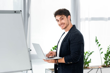 Fototapeta na wymiar handsome and smiling man in shirt holding laptop and looking at camera in office