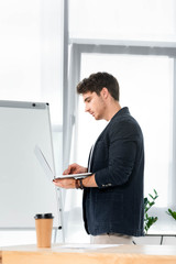 Fototapeta premium side view of handsome in shirt using laptop in office