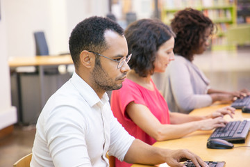 Serious adult male student studying in computer class. Row of man and women in casual sitting at table, using desktops, typing, looking at monitor. Online exam concept