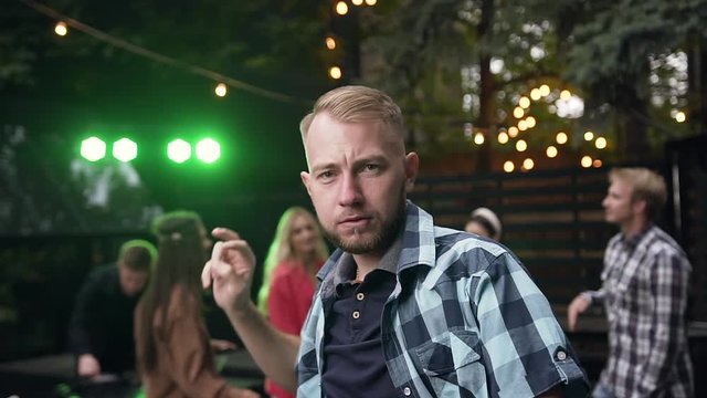 Close up of handsome bearded young man in stylish shirt rythmic dancing and making sign v on the background of group of mates