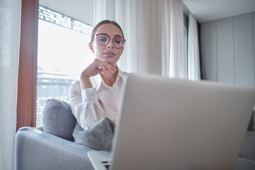 Enjoying time at home. Beautiful young smiling woman working on laptop and drinking coffee while sitting in a big comfortable chair at home