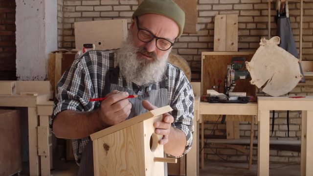 Lockdown of old trendy carpenter with beard and eyeglasses standing at workbench and making marks on wooden nesting box with pencil