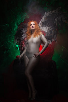 an evil tempting woman with large demon wings holds an Apple in a large cage and beckons to sin. Halloween photo plus size girl with red hair on a huge Gothic throne.