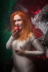Fototapeta premium Mysterious plus size red hair girl in skincolor beige outfit with black and white big demon wings on gothic cemetery. Witch or dark angel style. Woman tempts by apple in the middle of the night