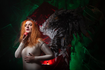 Mysterious plus size red hair girl in skincolor beige outfit with black and white big demon wings on gothic cemetery. Witch or dark angel style. Woman tempts by apple in the middle of the night