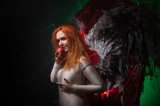 an evil tempting woman with large demon wings holds an Apple in a large cage and beckons to sin. Halloween photo plus size girl with red hair on a huge Gothic throne.