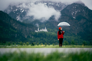 Asian woman hold umbrella standing in the meadow with background of green mountain and Neuschwanstein castle.  Concept of carefree, happy vacation.