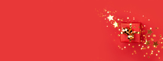 Banner with red gift box with golden bow on red background decorated golden stars confetti. Top view, minimal styled Christmas and holiday concept.