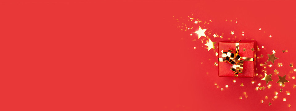 Banner with red gift box with golden bow on red background decorated golden stars confetti. Top view, minimal styled Christmas and holiday concept.