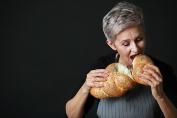 beautiful aged woman in apron biting bread on black background