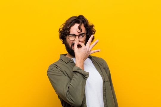 young crazy man feeling disgusted, holding nose to avoid smelling a foul and unpleasant stench against yellow wall