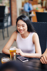 Happy Asian young woman sitting at the table and smiling while drinking beer in outdoor cafe