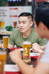 Asian young man drinking beer together with his friends while they sitting at the table in cafe