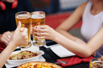 Close-up of young couple holding glasses with cold beer and toasting during their dinner at the table in cafe