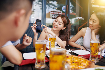 Asian young woman pointing at her mobile phone and telling her friends about good online store during their meeting in cafe