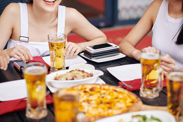 Close-up of young friends sitting at the table eating pizza and drinking beer with stack of mobile phones on the corner of the table