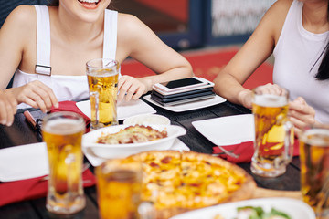 Close-up of young friends sitting at the table eating pizza and drinking beer with stack of mobile...