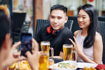 Asian young couple smiling and posing at camera of mobile phone while sitting at the table together with their friends in restaurant