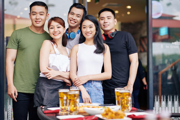 Portrait of Asian young friends smiling at camera while standing together near the table with food and drinks at the restaurant