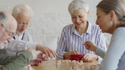 Tracking medium shot of group of four cheerful elderly people, two men and two women, having fun sitting at table and playing bingo game together in nursing home