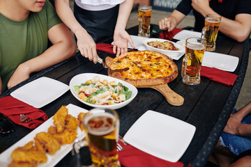 Close-up of young people sitting at wooden table eating pizza and salad and drinking beer during lunch at the restaurant