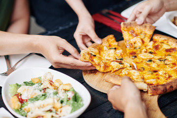 Close-up of people taking slices of pizza from the table and eating them during lunch in cafe