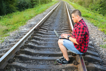 Guy with a laptop is sitting on the railway tracks. Freelance hipster worker. Man is working traveling.