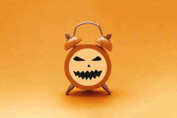 Halloween time concept. Orange alarm clock with evil jack lantern pumpkin face on orange background