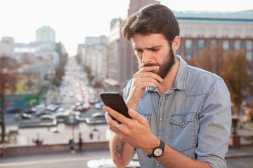 Young bearded man looking confused, browsing online on his smart phone, copy space. Handsome man...