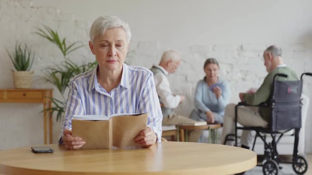 Rack focus of three elderly people, two men including disabled one and woman, playing cards in nursing home. Old woman with short grey hair reading book and thinking sadly sitting at table alone