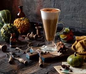 autumn traditional hot drink pumpkin spice latte in glass cup with foamy whipped cream on rustic wooden table with cinnamon, spoons and assorted mini pumpkins