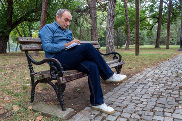 Middle Age Man Sitting on Wooden Bench in Park and Taking Notes