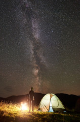 Back view of young man tourist resting at campfire beside camping and glowing tourist tent, enjoying view of night sky full of stars and Milky way. Vertical shot